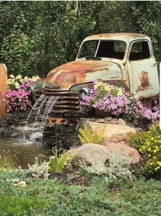 an old rusted truck is sitting in the middle of flowers and rocks near a pond