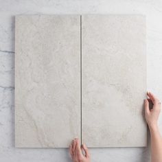 two hands are holding up a white marble wall panel with one hand and the other hand is pointing at it