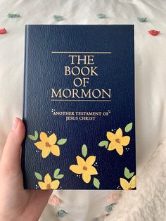 the book of mormon is being held up in front of a white bed with flowers on it