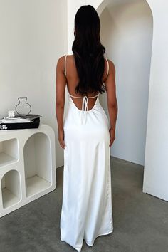 a woman in a white dress is standing near a shelf and looking at the wall