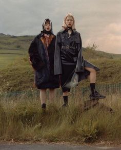 two women standing in tall grass with one wearing a black coat and the other a brown fur coat