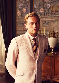 a man in a white suit and tie standing next to a table with an antique clock on it