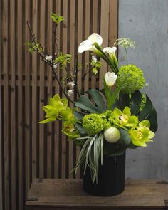 a black vase filled with green and white flowers on top of a wooden table next to a wall