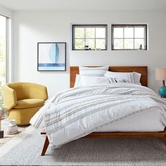 a bed with white sheets and pillows in a bedroom next to a chair, lamp and window