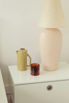 a white table topped with a lamp and a vase next to a coffee pot on top of a dresser