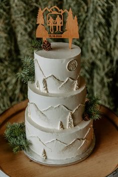 a three tiered white wedding cake with pine trees on the side and a wooden top