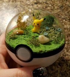 a hand holding a glass ball filled with green moss and small toy pokemon figurines