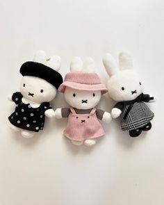 three small stuffed animals are sitting next to each other on a white surface, one is wearing a pink dress and the other has a black hat