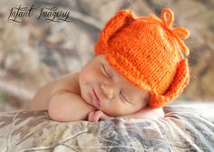 a baby wearing an orange knitted hat laying on top of a camouflage blanket with his eyes closed