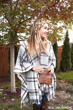 a woman standing in front of a tree wearing a plaid ponchet and holding a brown purse