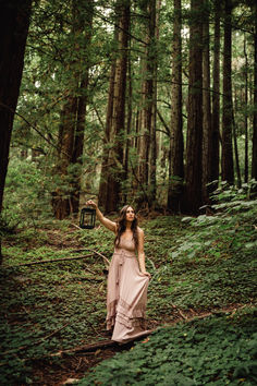 a woman in a long dress is walking through the woods with her arms outstretched up