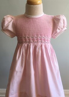 "This lovely dress is made up from a narrow pink and white stripe (like Oxford cloth stripes) in cotton or a cotton/poly blend. There is no fabric label. The manufacturer is \"Bon Tots by L & B\". The front bodice is entirely shirred and has double rows of hand embroidered white diamonds with rose pink and green leafed florals near waistline. The skirt is gathered and full and a longer length: mid-calf?? The sleeves are short and puffed with cuffs of white and narrow self piping at top. Neck Cute Striped Cotton Dress, Cotton Short Sleeve Dresses With Vertical Stripes, Short Sleeve Cotton Dress With Vertical Stripes, Cotton Dress With Vertical Stripes And Short Sleeves, Spring Cotton Dresses With Vertical Stripes, Spring Cotton Dress With Striped Hem, Pink Cotton Dress With Smocked Bodice, Fitted Spring Dress With Striped Hem, Spring Fitted Dress With Striped Hem