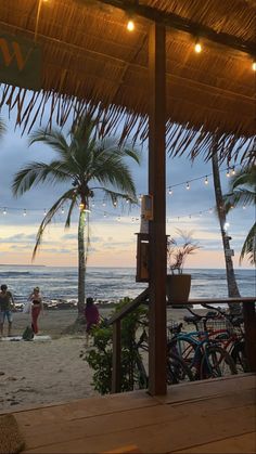 people are walking on the beach under a thatched roof with lights strung from it