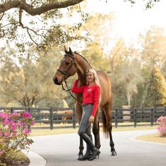 a woman standing next to a brown horse on top of a road with pink flowers