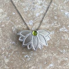 This handmade lotus flower pendant is hand-pierced and texturized from sterling silver. It features a smooth and clear, light green natural peridot gemstone. Peridot is the August birthstone. The pendant measures 1" in height by 1.6" in width and comes with your choice of a sterling silver chain necklace or an Omega necklace. The chain necklace is available in two sizes (16" or 20") and the Omega necklace is 16" with an attached 2" extender. The 16" chain necklace is shown on the model. • Handma Carter Jewelry, Fan Flower, Lotus Flower Pendant, Omega Necklace, Hand Piercing, August Birthstone, Sterling Silver Chain Necklace, Peridot Gemstone, Green Peridot