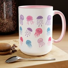 a pink and white coffee mug with jellyfish designs on it next to a spoon