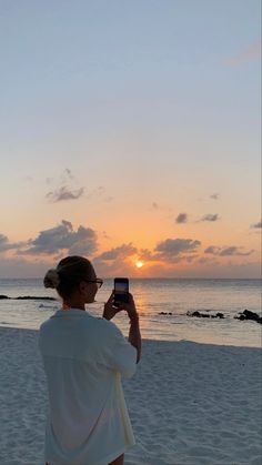 a woman sitting on the beach taking a photo with her cell phone at sunset or dawn