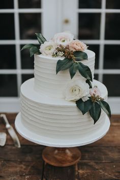 a white wedding cake with flowers and greenery on top, sitting on a wooden table