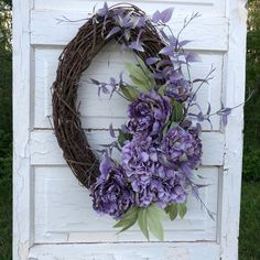 a wreath with purple flowers hanging on the side of a white door in front of some grass