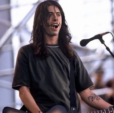 a man with long hair playing guitar and singing into a microphone at an outdoor concert