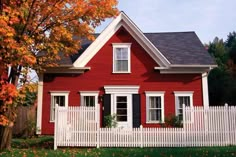 a red house with white picket fence and trees in the front yard on a fall day