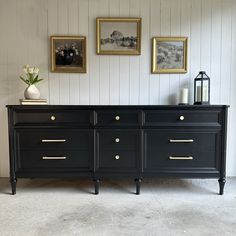 a black dresser with gold handles and knobs in a white walled room next to pictures on the wall