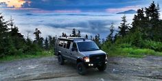 a truck parked on top of a dirt road in the middle of trees and clouds