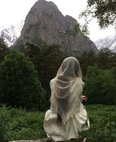 a veiled woman sitting on a log in front of a mountain with snow - capped peaks