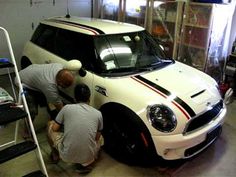 two men working on a white mini cooper in a garage with other people around the vehicle