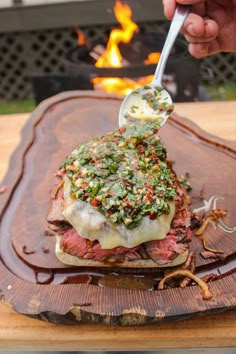 a person spooning sauce over a roast beef sandwich on a wooden platter with flames in the background