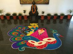 a colorfully painted peacock on the floor in front of a statue and potted plants