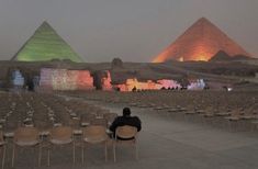 a man sitting on a chair in front of two pyramids with lights projected on them