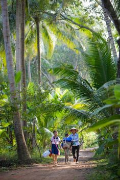 two people walking down a dirt path in the woods with bags on their back and one person holding a bike behind them