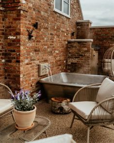 a bath tub sitting next to a brick wall near two wicker chairs and a potted plant