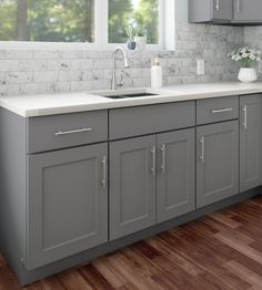 a kitchen with gray cabinets and white counter tops, wood floors and windows in the background