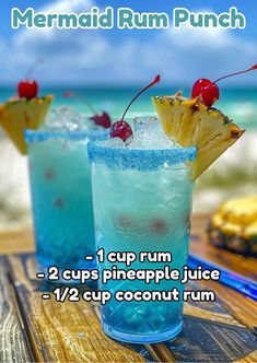 two blue cocktails with pineapple and cherries in them on a wooden table