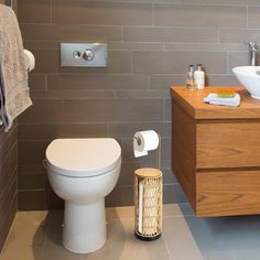 a white toilet sitting next to a wooden cabinet in a bathroom with gray tiles on the walls