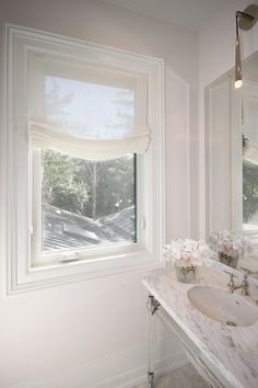 a bathroom with marble counter tops and white walls, along with two windows that have roman shades on them