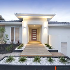 a modern house with white walls and wooden walkway leading to the front door, surrounded by plants