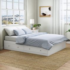 a white bed with blue sheets and pillows in a room next to a large window