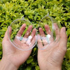 two hands holding small clear glass objects in front of green bushes