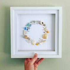 a hand is holding up a shadow box frame with sea shells and seashells in it