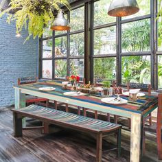 a dining room table and bench in front of large windows with potted plants on them