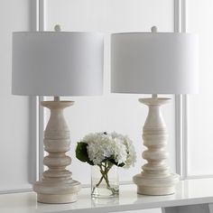 two white vases with flowers in them sitting on a table next to a lamp