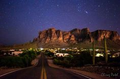 an empty road with mountains in the background and stars above it, at night time