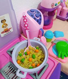 a pink toy stove top oven filled with pasta and veggies next to other toys