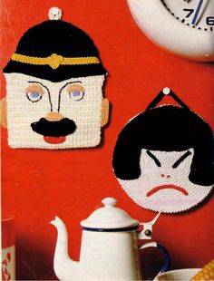 two crocheted pieces of cartoon characters on a red wall next to a teapot and clock