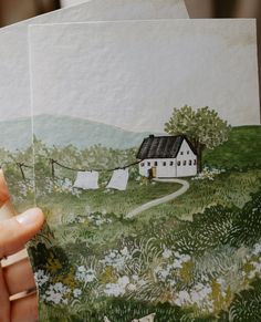 a person holding up some paper with a house on the hill in the back ground