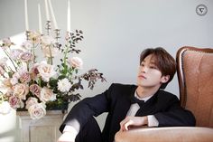 a young man in a tuxedo sitting on a chair next to a vase with flowers