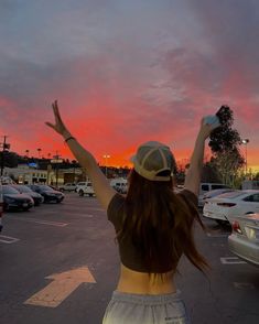 a woman is standing in a parking lot with her arms up to the sky as the sun sets